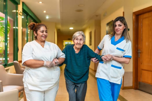 Two nurses are assisting an elderly woman as she walks down the hallway of a nursing home, providing support and care