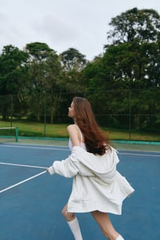 A young woman in a white oversized sweater joyful while playing tennis in a lush green outdoor setting, conveying energy and a sporty lifestyle, perfect for health and fitness themes