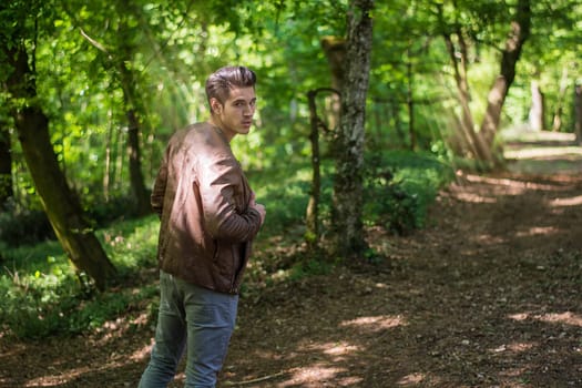 A young man in casual clothing strolls along a dirt path in a lush green forest. Sunlight filters through the trees, creating a serene atmosphere as he looks back over his shoulder.