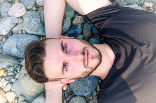 A young man lies back on smooth, round stones at a beach, enjoying the warm sun on a clear day. His relaxed expression conveys a sense of tranquility and peace with nature.