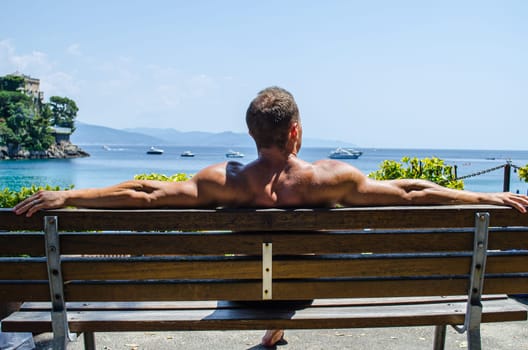 A man sits peacefully on a wooden bench, enjoying the warm sun and tranquil views of the ocean. Boats float gently on the water, creating a picturesque coastal atmosphere.