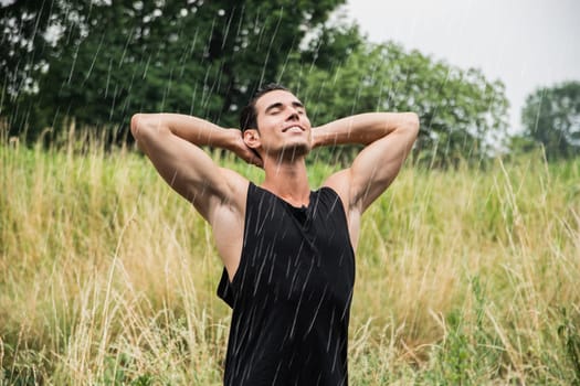 A man stands in a grassy field, arms raised and smiling, as rain falls around him. The lush greenery provides a vibrant backdrop on this warm summer day.