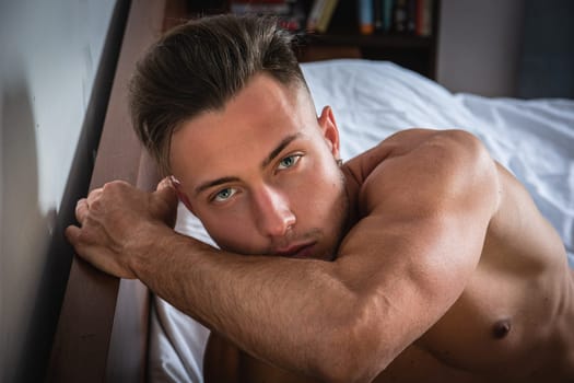 A young man lies on his side in bed, looking thoughtfully at the camera. Morning light gently illuminates his features, highlighting his well-defined arms and striking green eyes.