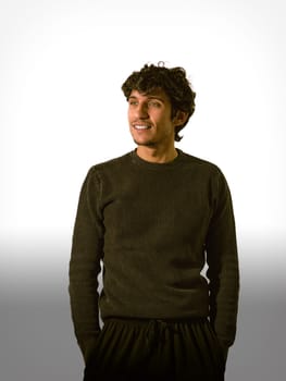A man with curly hair stands confidently, smiling softly while dressed in a dark sweater. Neutral tones in the background emphasize his relaxed demeanor in a casual indoor setting.