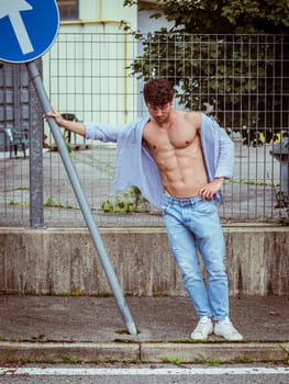 A young man stands confidently next to a street sign, wearing a light blue unbuttoned shirt and jeans. The setting appears urban, with greenery and modern buildings in the background.