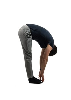 A man stands barefoot, bending forward to touch his toes. He wears a black shirt and grey pants, practicing flexibility and stretching in studio setting.