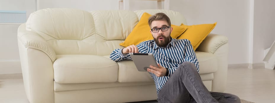 Young man lying on floor and using tablet device near sofa. Digital communication and casual home lifestyle concept. Banner copy space.