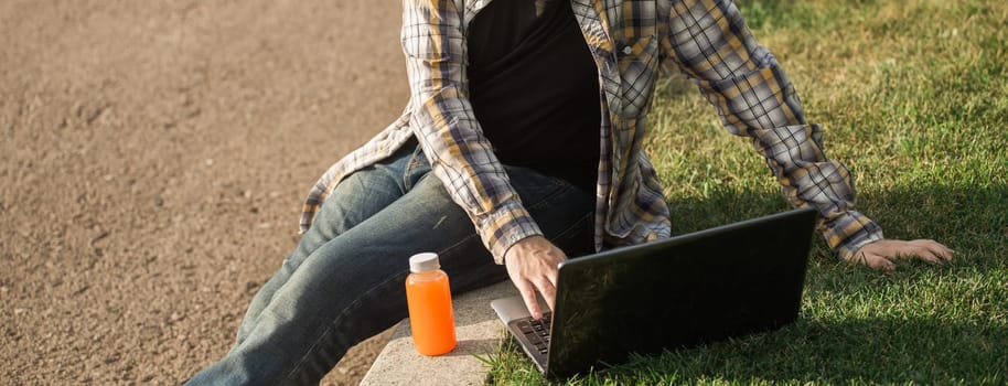 Man working outdoors on a laptop with juice beside him. Concept of remote work, productivity, and casual freelance lifestyle in urban parks. Banner with copy space