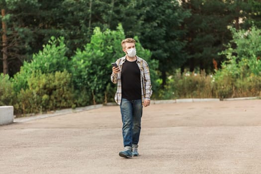 Man wearing a mask while walking in the park and checking his smartphone. Concept of modern life, health safety, and communication during pandemic times. Copy space.