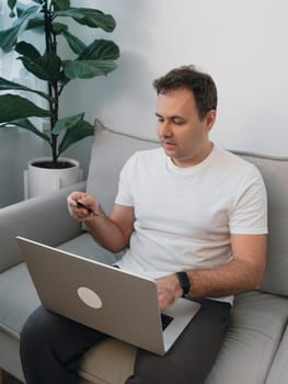 Side view of a man holding a bank card and verifying details in a modern living room with a fiddle leaf fig plant. Concept of secure online payment, digital banking, e-commerce and online shopping