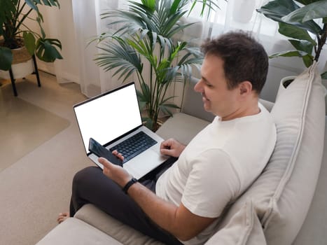 Smiling man sitting with laptop on knees, looking at smartphone. Laptop screen is visible, ideal for mockup use. Concept of positive digital interaction, remote work, and modern technology lifestyle.