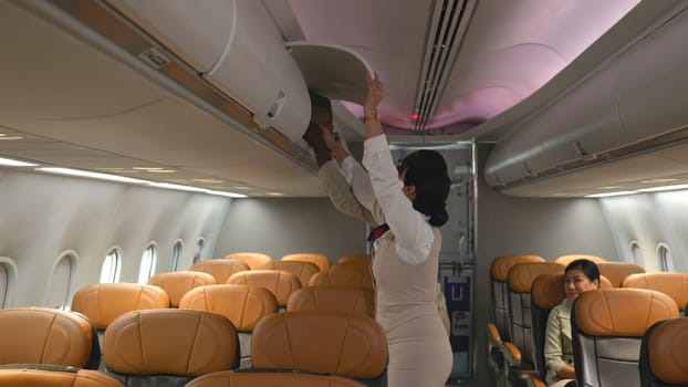 Friendly flight attendant. Air hostage helping passenger businessman put luggage on top shelf or cabin compartment on airplane, woman help black man lift suitcases putting overhead locker on airplane