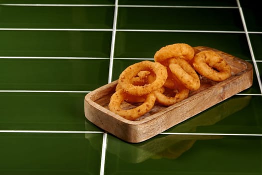 Delicious golden onion rings resting on a wooden tray, placed on a vibrant green tiled surface, creating a visually appealing culinary scene