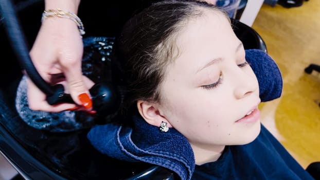 Client relaxes while having her hair washed in a salon basin, the stylist's hands gently massaging her scalp. Water ripples in the basin reflect the care of the salon experience, as a protective cape drapes over the client, ensuring comfort and neatness.