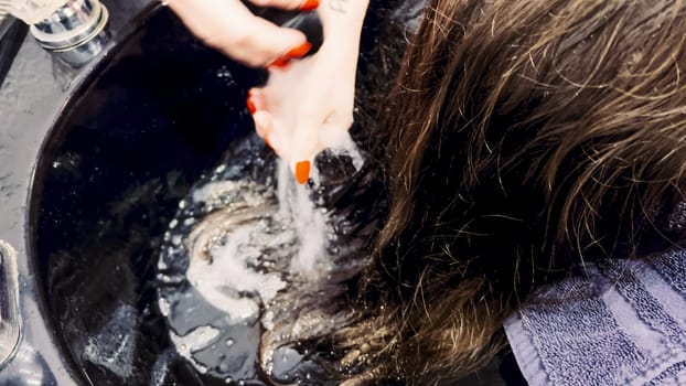 Client relaxes while having her hair washed in a salon basin, the stylist's hands gently massaging her scalp. Water ripples in the basin reflect the care of the salon experience, as a protective cape drapes over the client, ensuring comfort and neatness.