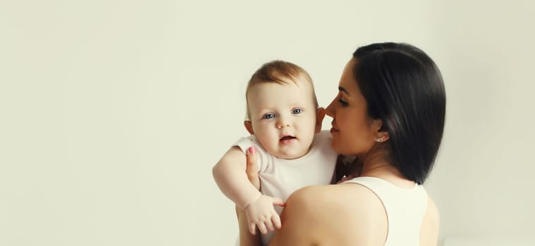 Happy young mother holding her cute baby sitting together in white room at home, studio background