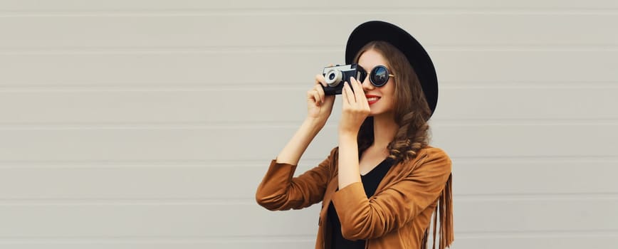 Portrait of beautiful happy young woman photographer taking picture with camera, stylish creative girl in black hat on gray background