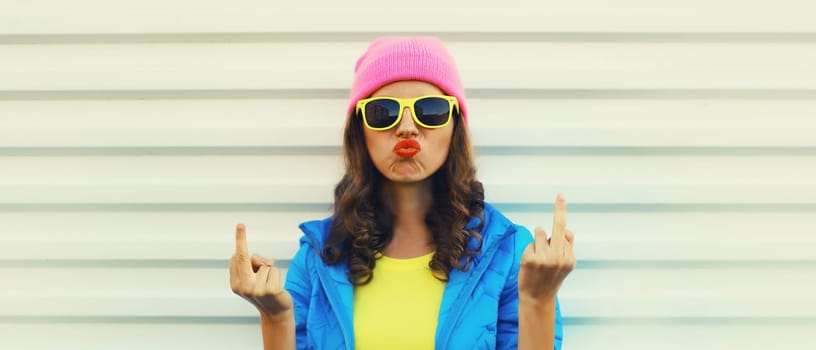 Portrait of bad angry young woman expression showing hand with middle finger sign, stylish girl wearing vivid colorful clothing, pink hat, glasses on white background