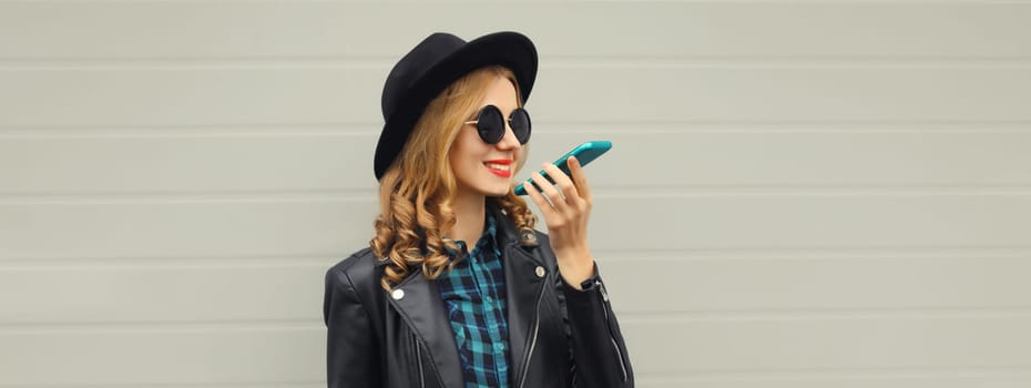 Beautiful happy smiling young woman calling on phone, stylish girl talking on the smartphone in black hat, jacket on gray background