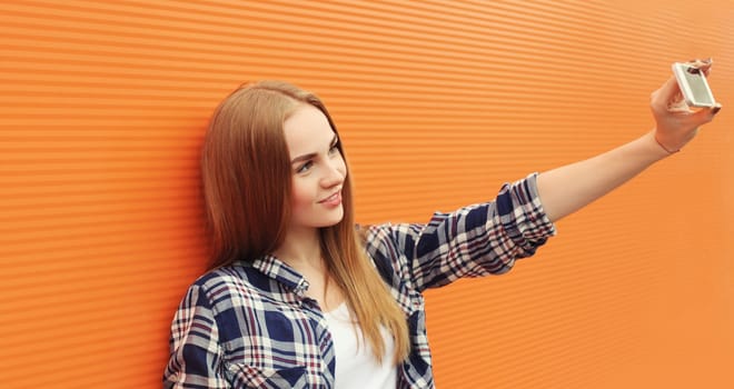 Summer portrait of happy smiling young woman taking selfie with phone, joyful girl holding smartphone on colorful orange background on city street