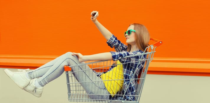 Stylish happy young woman taking selfie with phone, cool modern teenage girl having fun sitting in shopping trolley cart in the city on a summer day