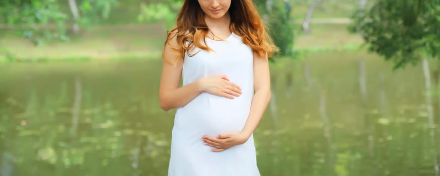 Close-up of happy young pregnant woman walking in summer park against lake background