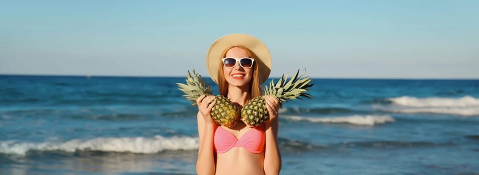 Summer vacation, beautiful happy smiling woman in pink bikini swimsuit, tourist straw hat holding pineapple fruits on the beach on sea coast background