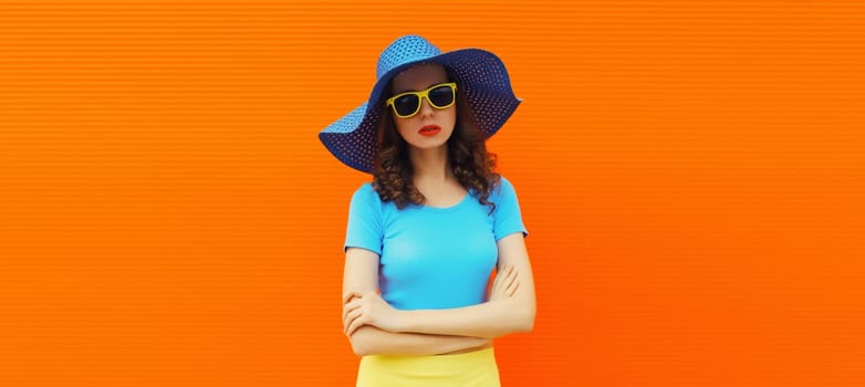 Beautiful stylish young woman, fashionable female model in summer blue straw hat posing on vivid orange wall background