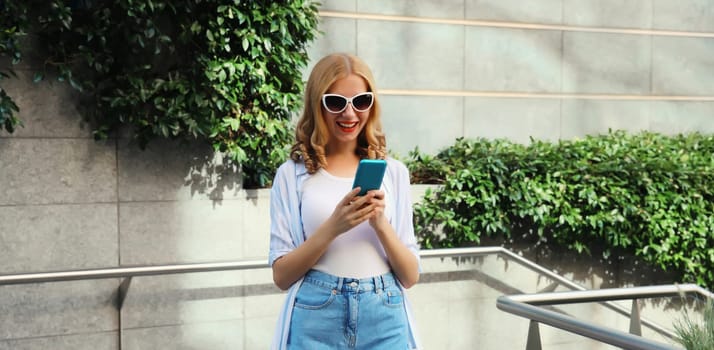 Stylish modern happy smiling young woman with phone standing in city park, girl holding smartphone on street, urban environment background with green plants