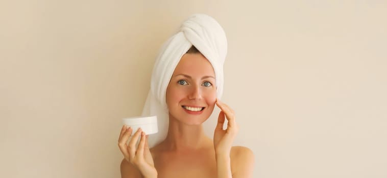 Natural beauty portrait of happy smiling young caucasian woman touches her clean skin applying face cream while drying wet hair with white bath towel on her head