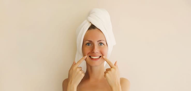 Portrait of happy smiling caucasian young woman points to her white clean teeth while drying her hair with towel on her head after shower in the morning at home