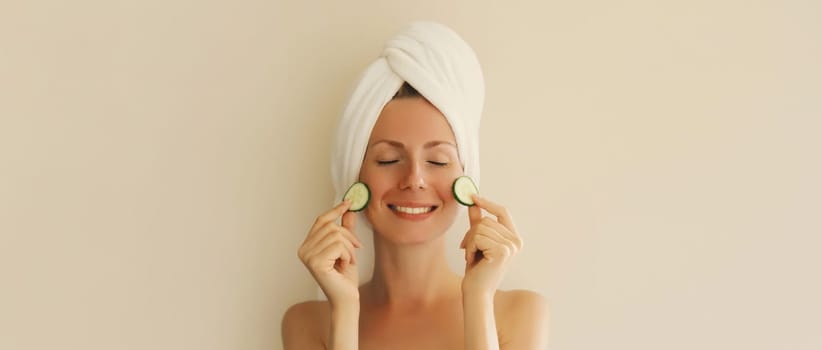 Natural beauty portrait of happy smiling young caucasian woman covers her clean skin holds slices of cucumber drying hair with white bath towel on her head at home