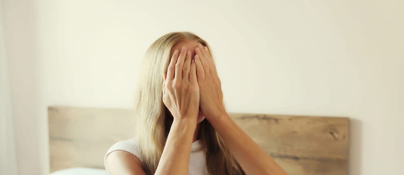 Upset sad crying young woman covering her eyes and face with her hands while experiencing depression, stress, fatigue, mental suffering after sleeping sitting on the bed at home