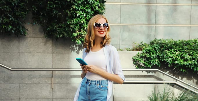 Stylish modern happy smiling young woman with phone standing in city park, girl holding smartphone on street, urban wall background with green plants