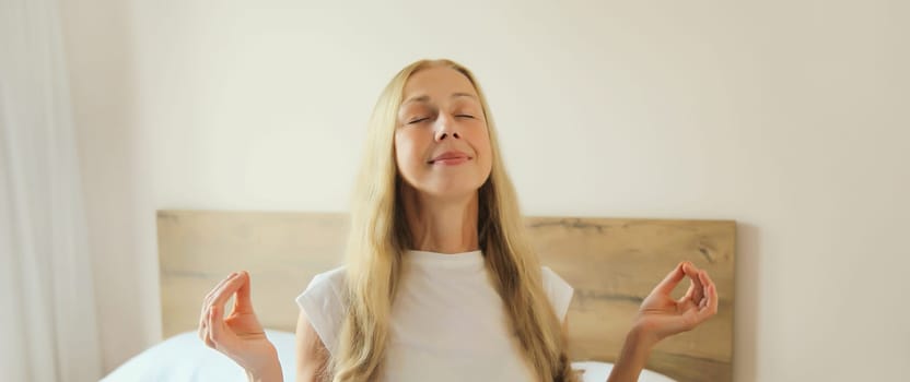 Calm relaxed healthy young woman meditates, girl practicing doing yoga exercises sitting in lotus pose on white bed at home, stress relief