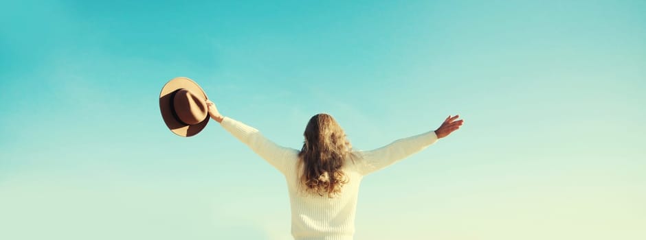 Back view of happy young woman raising her hands up on sunny beach, enjoying amazing landscape, warm weather, wear hat, white sweater, standing on sea coast, nature background