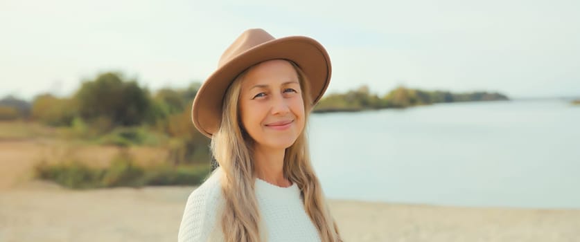 Stylish happy woman standing on sunny beach, warm weather, smiles, wear hat, white sweater, on sea coast background