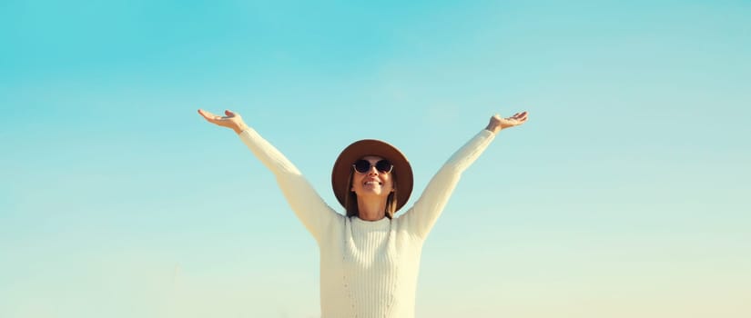 Happy smiling woman traveler raising her hands up on beach, enjoying warm weather, wear hat, white sweater, standing on sea coast, blue sky, nature background