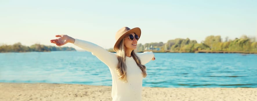 Happy smiling woman traveler raising her hands up on beach, enjoying warm weather, wear hat, white sweater, standing on sea coast, blue sky, nature background