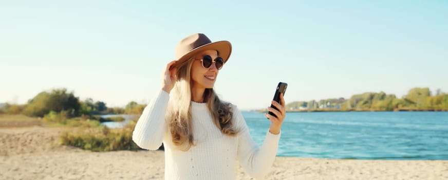 Travel, adventure, stylish happy woman taking selfie with phone on sunny beach, girl using smartphone in hat, white sweater standing on sea or river coast landscape, nature background