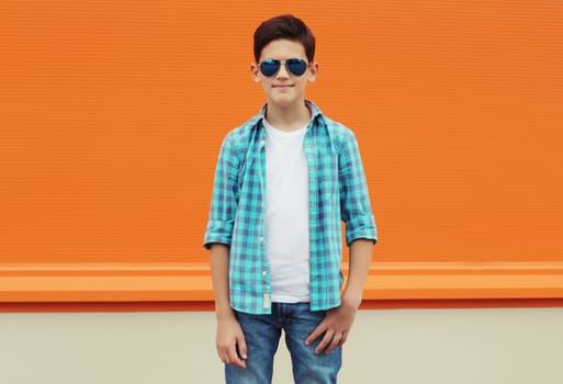 Modern teenage kid boy in casual clothes with glasses standing on city street, orange wall background