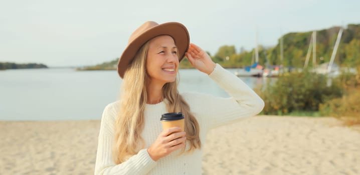 Stylish happy woman with cup of coffee drink standing on beach, smiles, looking away, wear hat, white sweater, on sea coast background