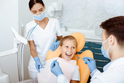 Fast, painless and modern examination and treatment of oral cavity. Smiling little girl opens her mouth in medical chair and looks at doctor in white coat, protective mask and rubber gloves with nurse