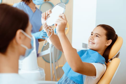 Good job and healthy teeth. Happy girl in dental chair with napkin looks in mirror in interior office with doctors in protective masks and uniforms looks at patient after treatment, free space