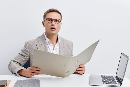 Young professional man with glasses, dressed in a gray checked suit, displaying shock or confusion while examining a big paper document at a clean, modern office desk with a laptop and notepad. Concept: workplace stress or unexpected business findings.