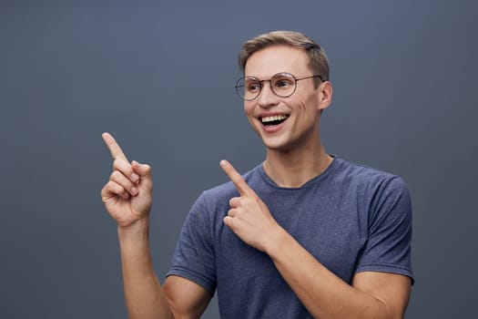 smiling young caucasian man in glasses and blue t-shirt point fingers to side and look excited holding mobile phone in hand isolated on gray background, man using chat AI technology for digital communication concept
