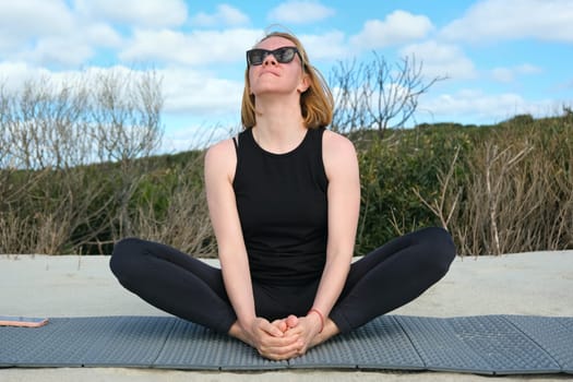 Woman practicing yoga outdoors, sitting in butterfly pose on a mat, enjoying fresh air, natural surroundings, and blue sky, embracing mindfulness and relaxation.
