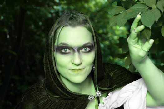 A woman dressed as a green forest spirit or witch looks out of the bushes