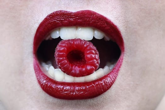 A woman with red lips holds a raspberry between her teeth