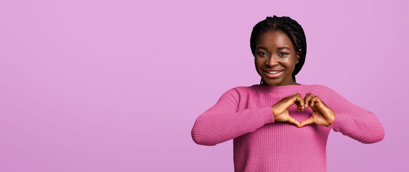 Romantic black lady shaping hands like heart, making love gesture near chest, affectionate african woman expressing kindness, smiling at camera, standing over purple studio background , copy space